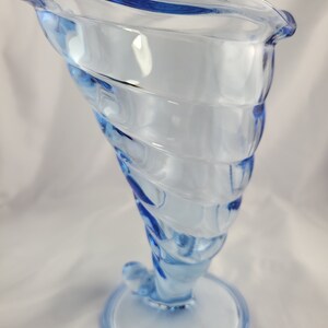 May include: A light blue glass vase with a swirling design. The vase has a wide mouth and a narrow base.