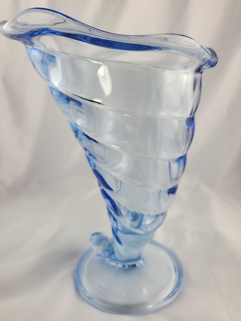 May include: A light blue glass vase with a swirling design. The vase has a wide mouth and a narrow base.