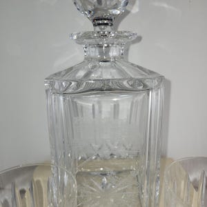 May include: A clear glass decanter with a faceted stopper and four matching glasses. The decanter has a square body with a cut glass design.