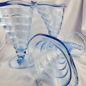 May include: Three blue glass ice cream dishes with a swirl design. The dishes are made of clear glass with a blue tint and have a scalloped rim.