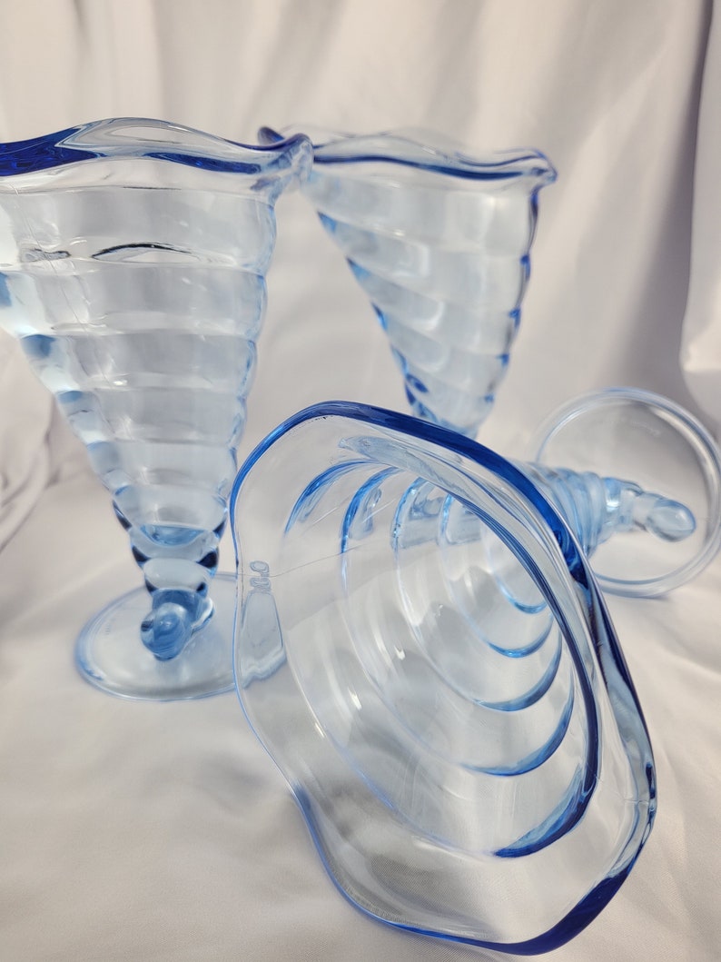 May include: Three blue glass ice cream dishes with a swirl design. The dishes are made of clear glass with a blue tint and have a scalloped rim.