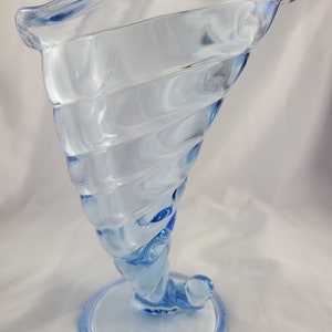 May include: A light blue glass vase with a swirling design. The vase has a wide mouth and a round base.