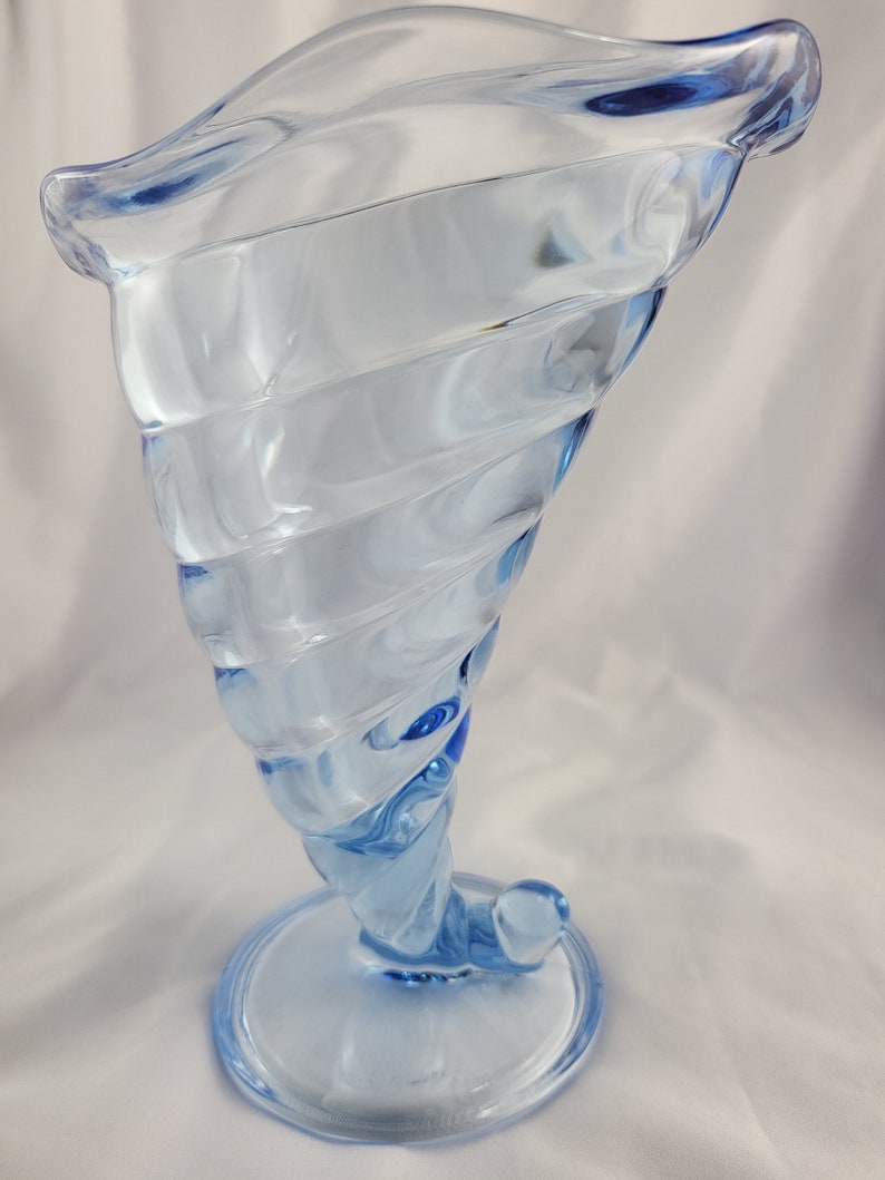 May include: A light blue glass vase with a swirling design. The vase has a wide mouth and a round base.