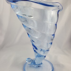 May include: A light blue glass vase with a swirling design. The vase has a wide mouth and a narrow base.