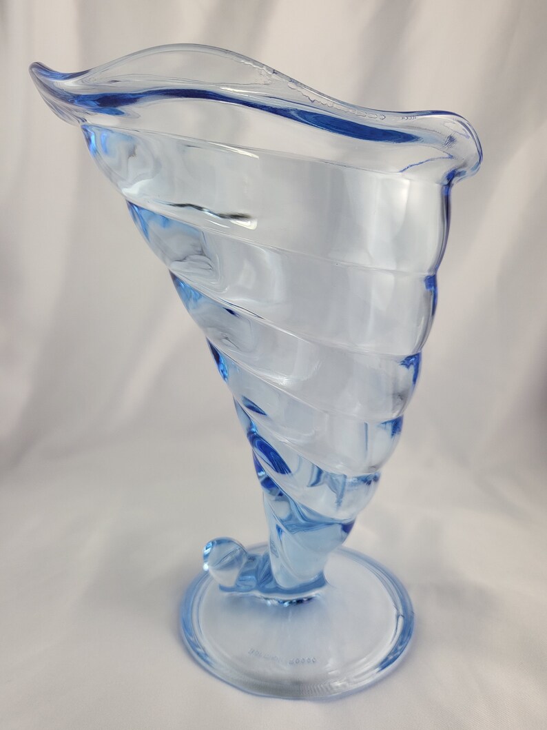 May include: A light blue glass vase with a swirling design. The vase has a wide mouth and a narrow base.