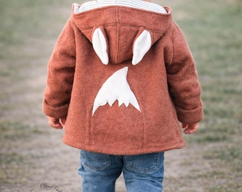 Könnte beinhalten: Eine rostfarbene Kapuzenjacke mit weißen Stoffohren an der Kapuze und einem weißen, dreieckigen Aufnäher auf dem Rücken. Die Jacke hat ein gestreiftes Innenfutter in der Kapuze und wird mit einer blauen Jeans und beigen Schuhen getragen.
