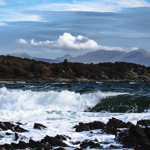 Może przedstawiać: Dramatyczny morski krajobraz z dużą falą rozbijającą się o skaliste brzegi. Woda jest ciemnoniebieska, a niebo jasnoniebieskie z białymi chmurami. W oddali widać pasmo górskie.