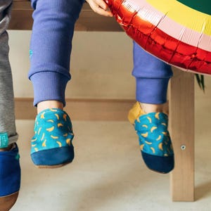 Puede incluir: Calzado infantil en varios estilos. Un par presenta un diseño azul y marrón, otro tiene un estampado de plátanos amarillos sobre fondo turquesa, y un tercer par tiene un estampado crema y rojo. Los niños llevan pantalones grises y azules.