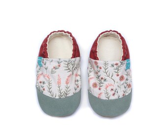 Può includere: Un paio di scarpine per neonati con suola morbida, caratterizzate da una tomaia in tessuto bianco con un delicato motivo floreale nei toni del rosa e del verde. Le scarpe presentano una punta in camoscio verde salvia, un colletto in tessuto rosso scuro e una piccola etichetta laterale color ottanio.