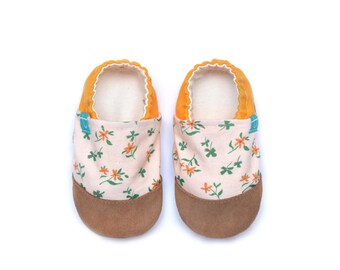 Può includere: Un paio di scarpine per bambini con motivo floreale. Le scarpe hanno una suola in pelle scamosciata marrone chiaro, una tomaia color crema con fiori arancioni e foglie verdi e un bordo arancione. L'interno è bianco.