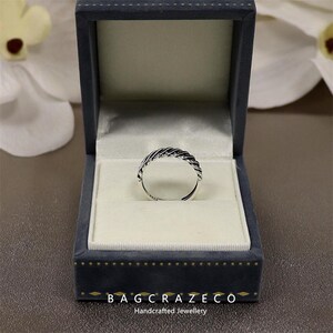May include: A silver braided ring presented in a blue velvet jewellery box. The ring's woven pattern is the focal point, set against a white interior. The open box is on a brown surface. The text "BAG CRAZECO Handcrafted Jewellery" is visible.