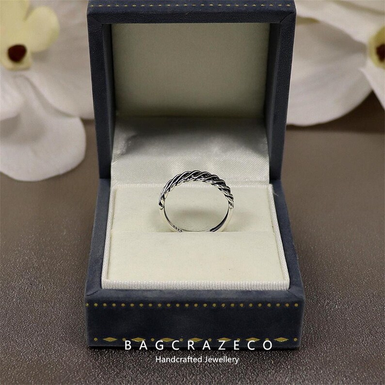 May include: A silver braided ring presented in a blue velvet jewellery box. The ring's woven pattern is the focal point, set against a white interior. The open box is on a brown surface. The text "BAG CRAZECO Handcrafted Jewellery" is visible.