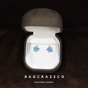 May include: Flower-shaped earrings with blue and silver detailing, presented in an open brown jewellery box with a white interior. The earrings are the central focus, set against a dark brown backdrop. The text "BAGCRAZECO" and "Handcrafted Jewellery" are at the bottom.