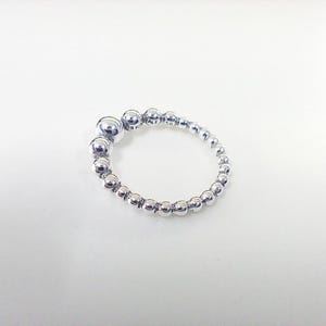 May include: A silver ring featuring a series of small, round beads. The ring has a unique design with larger beads clustered together at the top, gradually decreasing in size as they encircle the finger. The ring is set against a white background.