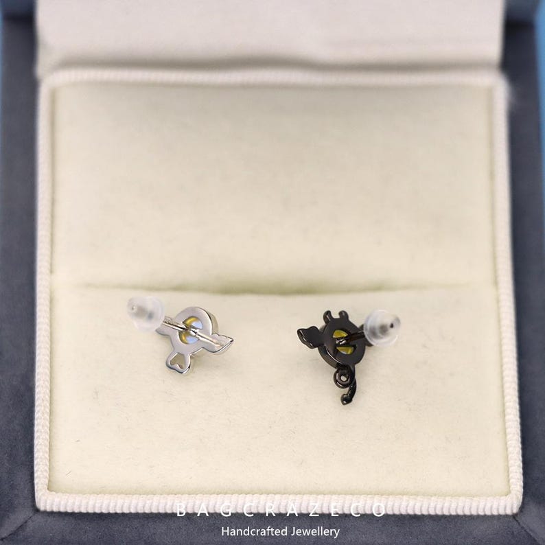 May include: A pair of earrings in a white box. One earring is silver with a yellow center and the other is black with a yellow center. The earrings are in the shape of a musical note.