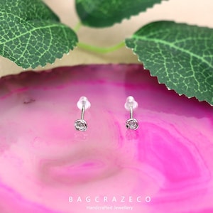 May include: A pair of silver rose stud earrings on a pink and white background. The earrings are small and delicate, with a simple rose design.