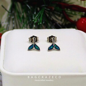 May include: A pair of silver stud earrings with a mermaid tail design. The tails are filled with a blue-green iridescent material. The earrings are displayed on a white surface with a blurred background of greenery and a red bauble.