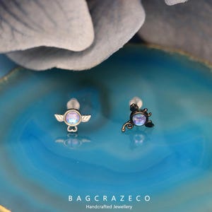 May include: Two silver and black body jewelry pieces with iridescent blue gems. The piece on the left has a small angel wing design and the piece on the right has a small scorpion design.