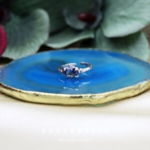 May include: A silver ring with a purple gemstone set in a textured band. The ring is displayed on a blue and gold agate coaster.