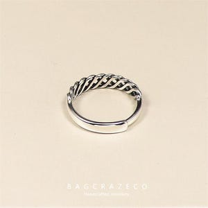 May include: A silver ring featuring a woven design across the top. The ring has a smooth band and a textured, braided pattern. The ring is a piece of handcrafted jewellery.