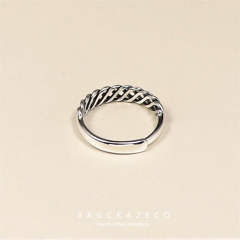 May include: A silver ring featuring a woven design across the top. The ring has a smooth band and a textured, braided pattern. The ring is a piece of handcrafted jewellery.