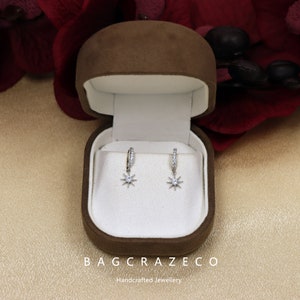 May include: A pair of silver star-shaped dangle earrings with clear cubic zirconia stones, displayed in a brown velvet jewelry box. The box has the text "Bagcrazeco Handcrafted Jewellery" on the bottom.