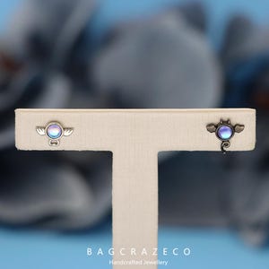 May include: A pair of earrings featuring a small, iridescent blue and purple gemstone. One earring is shaped like a bat with wings, while the other is shaped like an angel with wings. Both earrings are on a white display stand.