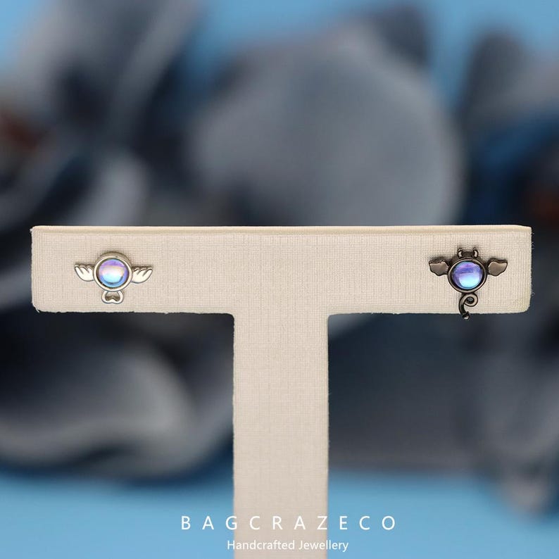 May include: A pair of earrings featuring a small, iridescent blue and purple gemstone. One earring is shaped like a bat with wings, while the other is shaped like an angel with wings. Both earrings are on a white display stand.