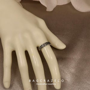 May include: A silver ring with a woven pattern is shown on a mannequin hand. The ring has a series of parallel lines, giving it a textured look. The ring is a piece of jewellery. The text "BAG CRAZECO Handcrafted Jewellery" is visible at the bottom.