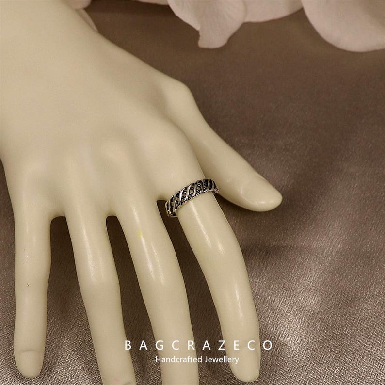May include: A silver ring with a woven pattern is shown on a mannequin hand. The ring has a series of parallel lines, giving it a textured look. The ring is a piece of jewellery. The text "BAG CRAZECO Handcrafted Jewellery" is visible at the bottom.