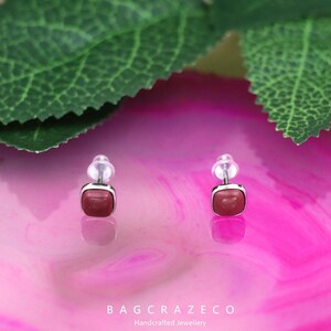 May include: A pair of silver stud earrings with square red gemstone accents. The earrings are displayed on a pink and white background.