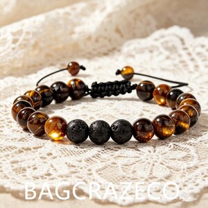 Puede incluir: Una pulsera de cuentas con piedras de ojo de tigre marrón pulidas y tres cuentas de lava negras mate y porosas en el centro. La pulsera está ensartada en un cordón negro con un cierre de nudo deslizante ajustable y dos cuentas en los extremos.