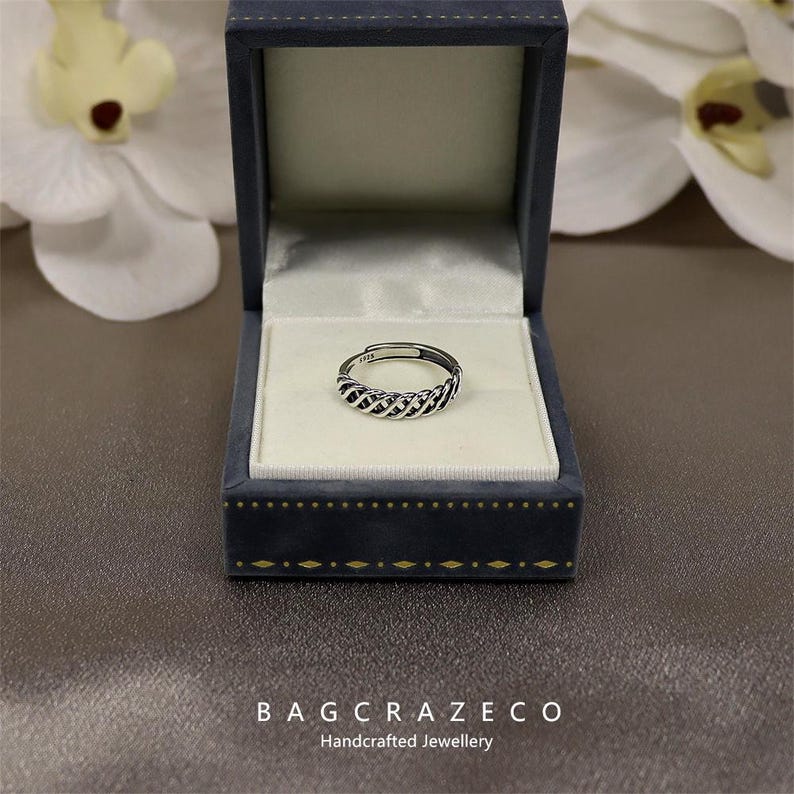 May include: A silver ring with a woven pattern, presented in a dark blue jewellery box with a white interior. The ring is adjustable. The box is on a brown surface, with white flowers in the background. The text "BAG CRAZECO Handcrafted Jewellery" is at the bottom.