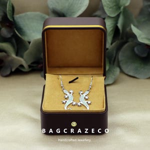 Puede incluir: Pendientes de plata con forma de lagarto, con un diseño blanco y azul claro, presentados en una caja de joyería marrón abierta con interior dorado. El texto "BAGCRAZECO Handcrafted Jewellery" es visible en la parte inferior.