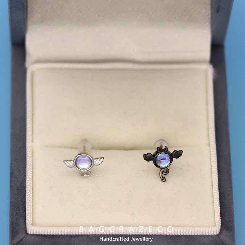 May include: Two silver and black body jewelry pieces with iridescent stones and wings. The jewelry is displayed in a white velvet box with the text "BAGGRAZECO Handcrafted Jewellery".