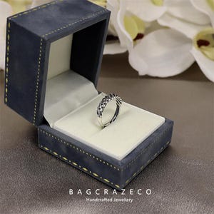 May include: A silver ring with a woven design sits in an open, dark blue velvet jewellery box. The box has a white satin interior and gold stitching. The text "BAG CRAZECO Handcrafted Jewellery" is visible on the box.