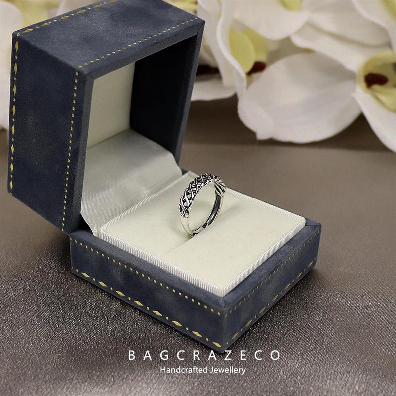 May include: A silver ring with a woven design sits in an open, dark blue velvet jewellery box. The box has a white satin interior and gold stitching. The text "BAG CRAZECO Handcrafted Jewellery" is visible on the box.