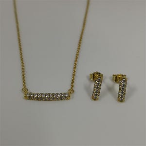 May include: A gold-toned jewelry set featuring a necklace and matching earrings. The necklace has a delicate chain with a horizontal bar pendant encrusted with small, clear gemstones. The earrings are small, vertical bars with similar gemstone detailing.