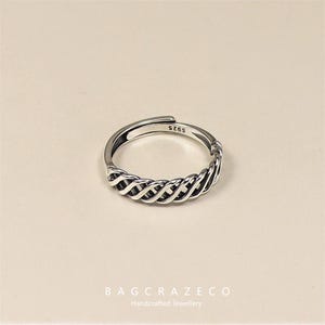 May include: A silver adjustable ring with a woven design. The ring has a braided pattern around the band and is stamped with "S925". The ring is displayed on a neutral background. The brand name "BAGCRAZECO" and "Handcrafted Jewellery" are at the bottom.