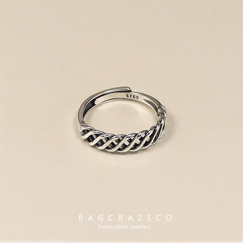 May include: A silver adjustable ring with a woven design. The ring has a braided pattern around the band and is stamped with "S925". The ring is displayed on a neutral background. The brand name "BAGCRAZECO" and "Handcrafted Jewellery" are at the bottom.