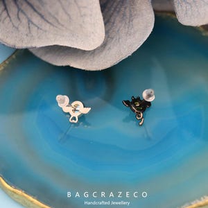 May include: A pair of earrings with a black and a silver finish. The black earring features a small, detailed design of a heart with wings. The silver earring features a small, detailed design of a heart with wings. The earrings are on a blue background.