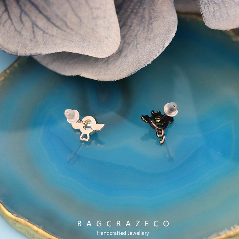 May include: A pair of earrings with a black and a silver finish. The black earring features a small, detailed design of a heart with wings. The silver earring features a small, detailed design of a heart with wings. The earrings are on a blue background.