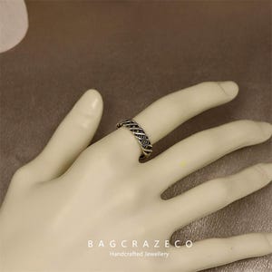 May include: A silver ring with a woven design is shown on a hand. The ring has a textured appearance, with a pattern of interwoven metal strands. The ring is a piece of handcrafted jewellery.