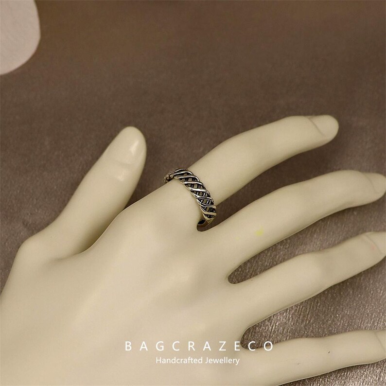 May include: A silver ring with a woven design is shown on a hand. The ring has a textured appearance, with a pattern of interwoven metal strands. The ring is a piece of handcrafted jewellery.