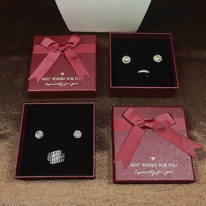 May include: Four red gift boxes with matching bows, each containing jewellery. Two boxes hold earrings, one with emerald accents and the other with diamond-like studs. The other two boxes contain a ring. Text on the boxes reads "BEST WISHES FOR YG" and "Especially for you."