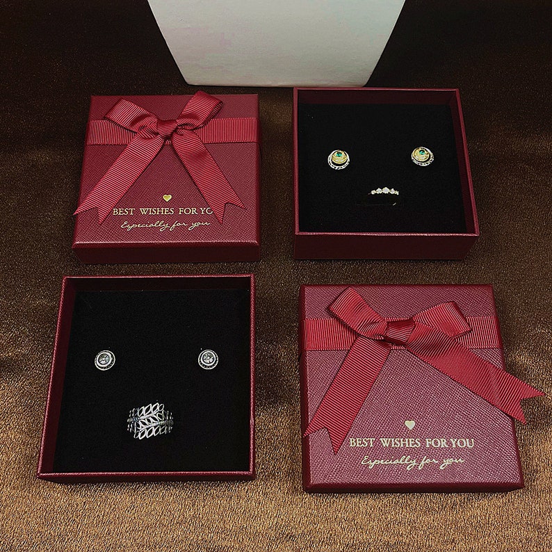 May include: Four red gift boxes with matching bows, each containing jewellery. Two boxes hold earrings, one with emerald accents and the other with diamond-like studs. The other two boxes contain a ring. Text on the boxes reads "BEST WISHES FOR YG" and "Especially for you."