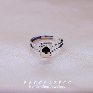 Può includere: Un anello d'argento con una doppia fascia che presenta un asso di picche nero con la scritta "BICYCLE" stampata su di esso.