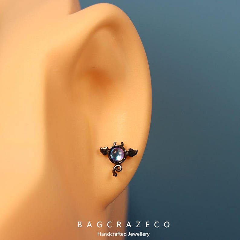 May include: A black bat with iridescent purple wings and a round gem in the center. The bat is attached to a small, curved tail. The bat is a body piercing jewelry piece.