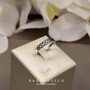 May include: A silver ring with a woven pattern is presented on a beige jewellery stand. The ring's intricate design is highlighted against a backdrop of blurred white flowers. The text "BAG CRAZECO Handcrafted Jewellery" is at the bottom.