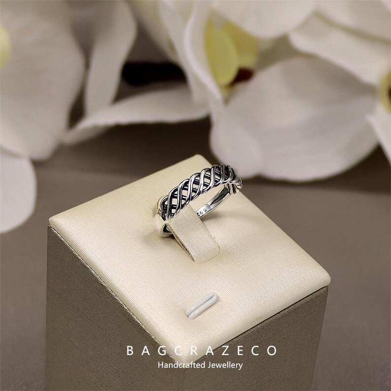 May include: A silver ring with a woven pattern is presented on a beige jewellery stand. The ring's intricate design is highlighted against a backdrop of blurred white flowers. The text "BAG CRAZECO Handcrafted Jewellery" is at the bottom.